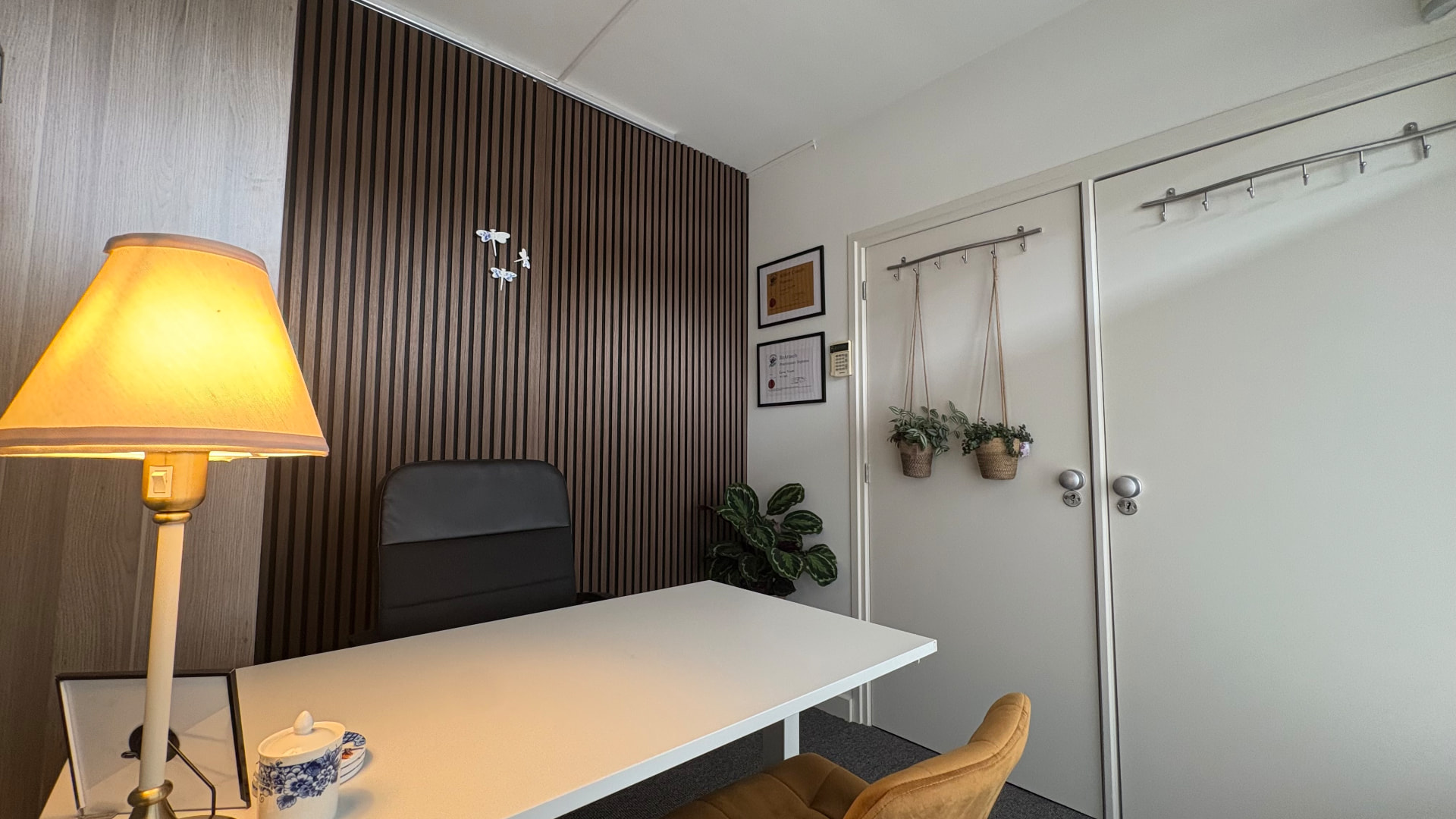 Behandelkamer van Praktijk Sluijter in Vlijmen: wit bureau met okergele stoel tegen houten lamellenwand